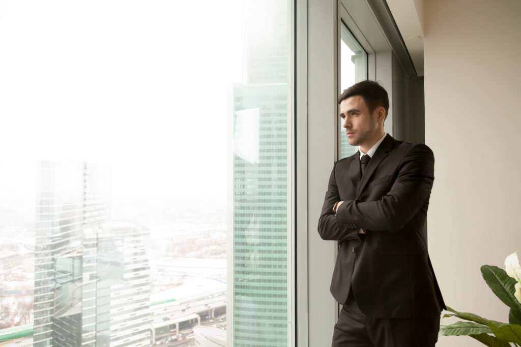 Pensive businessman standing near large window in office and looking on urban landscape with skyscrapers outside. Serious business leader pondering difficult decision, thinking about future challenges