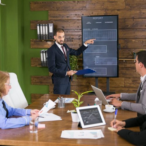 Businessman in suit pointing at charts on tv screen in a meeting with his colleagues. Leadership meeting in conference room.